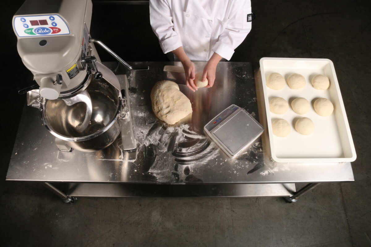 looking down at a prep table as a chef prepares breads with flour and a globe mixer